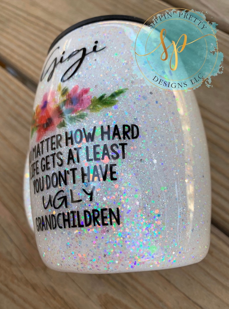 Puede incluir: Vaso blanco brillante con la palabra "Gigi" en escritura negra, detalles florales y el texto "No importa lo dif&iacute;cil que se ponga la vida, al menos no tienes nietos feos". Se ve un logotipo de "Sippin Pretty Designs LLC".
