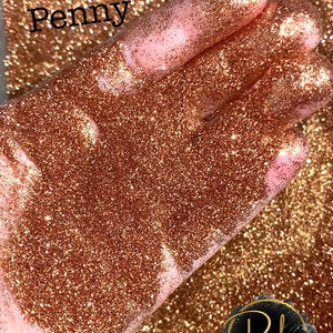 May include: A close-up of a copper-colored glitter, with a hand holding a small amount of the glitter. The glitter is labeled "Penny" and the image is watermarked with "Posh Glitter, LLC."