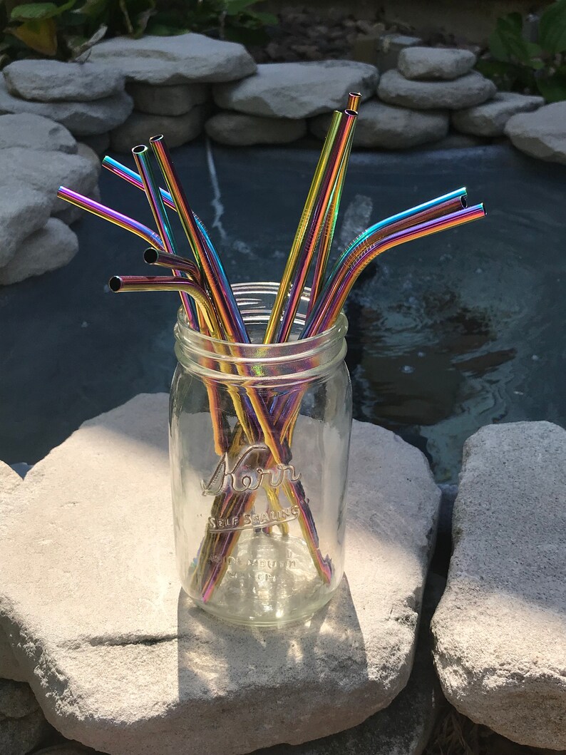 May include: A glass jar filled with a bundle of rainbow colored metal drinking straws. The jar is sitting on a gray rock.
