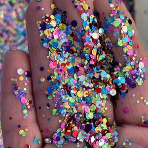 May include: Close-up of a hand holding a vibrant mix of colorful, round glitter. The glitter pieces vary in size and feature a rainbow of hues, including pink, blue, green, yellow, and purple. The glitter is scattered across the fingers.