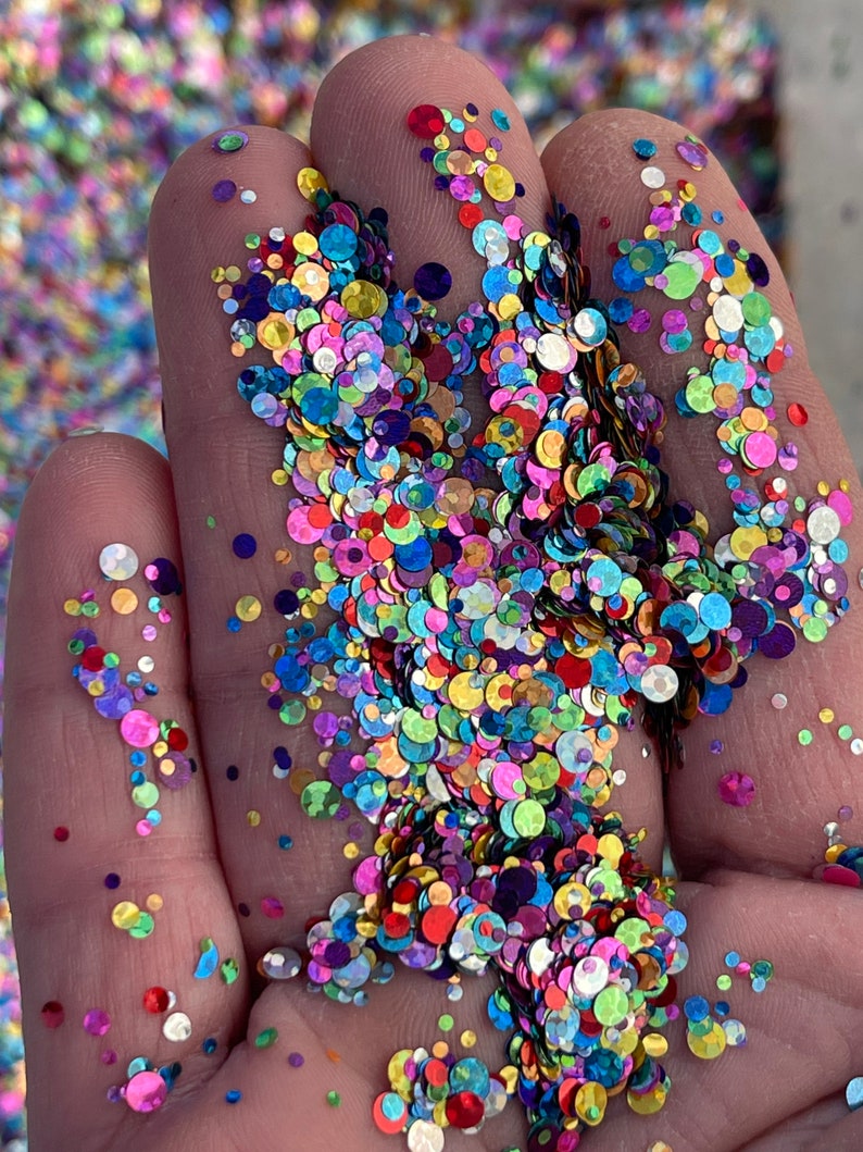 May include: Close-up of a hand holding a vibrant mix of colorful, round glitter. The glitter pieces vary in size and feature a rainbow of hues, including pink, blue, green, yellow, and purple. The glitter is scattered across the fingers.