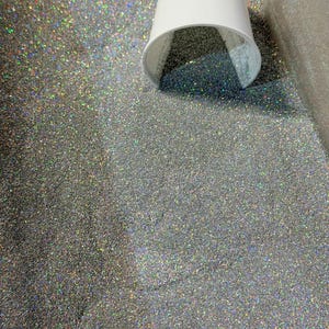 May include: A white paper cup is tipped over, spilling silver glitter onto a surface. The glitter is densely packed and reflects multiple colors. The background is a light gray color.