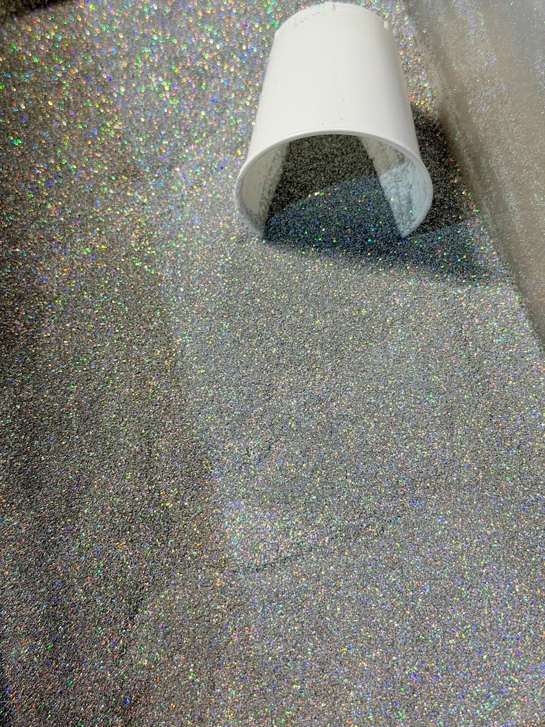 May include: A white paper cup is tipped over, spilling silver glitter onto a surface. The glitter is densely packed and reflects multiple colors. The background is a light gray color.