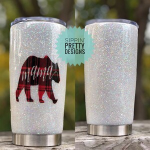 Puede incluir: Un vaso blanco con purpurina y base de acero inoxidable. El vaso presenta un dise&ntilde;o de oso a cuadros rojos y negros con la palabra "mama" en escritura cursiva. Tambi&eacute;n se ve el texto "Sippin' Pretty Designs".