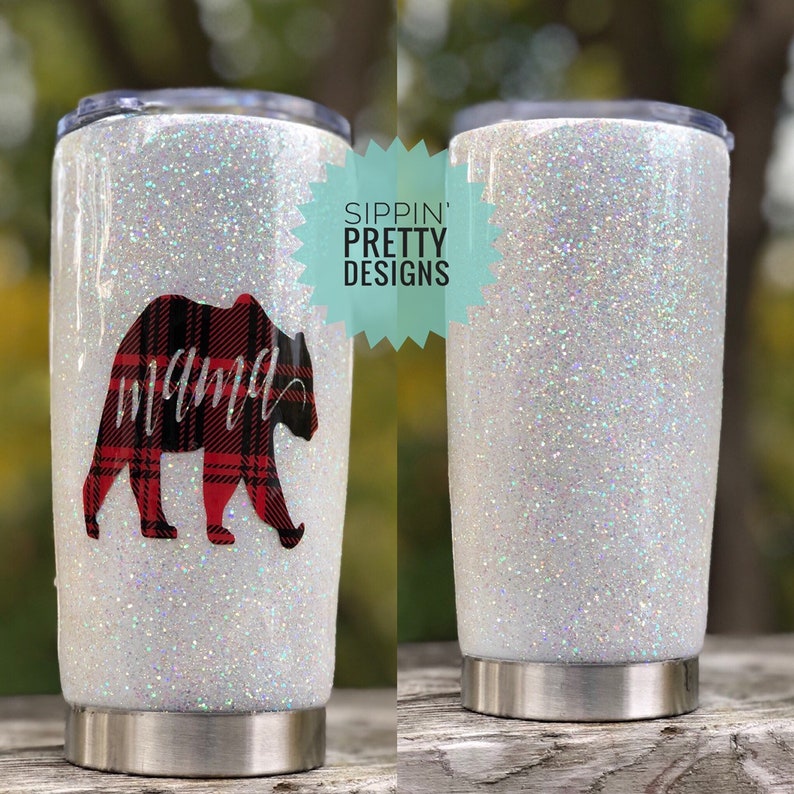 Puede incluir: Un vaso blanco con purpurina y base de acero inoxidable. El vaso presenta un dise&ntilde;o de oso a cuadros rojos y negros con la palabra "mama" en escritura cursiva. Tambi&eacute;n se ve el texto "Sippin' Pretty Designs".