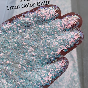 May include: A close-up of a pile of iridescent white glitter with a slight blue hue. The glitter is 1mm in size and has a color shift effect. The text "Pearl 1mm Color Shift" is visible in the image.