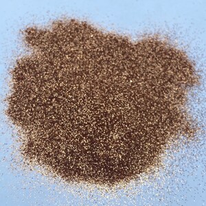 May include: A close-up of a pile of copper-colored glitter on a light blue background. The word "PENNY" is written in brown letters at the top of the image.
