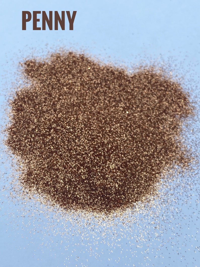 May include: A close-up of a pile of copper-colored glitter on a light blue background. The word "PENNY" is written in brown letters at the top of the image.