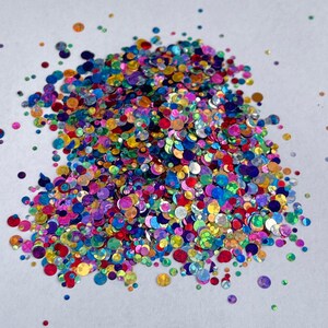 May include: A pile of colorful, iridescent glitter in various sizes and shapes. The glitter includes shades of red, blue, green, purple, and gold. The glitter is scattered on a white surface, creating a sparkling effect.