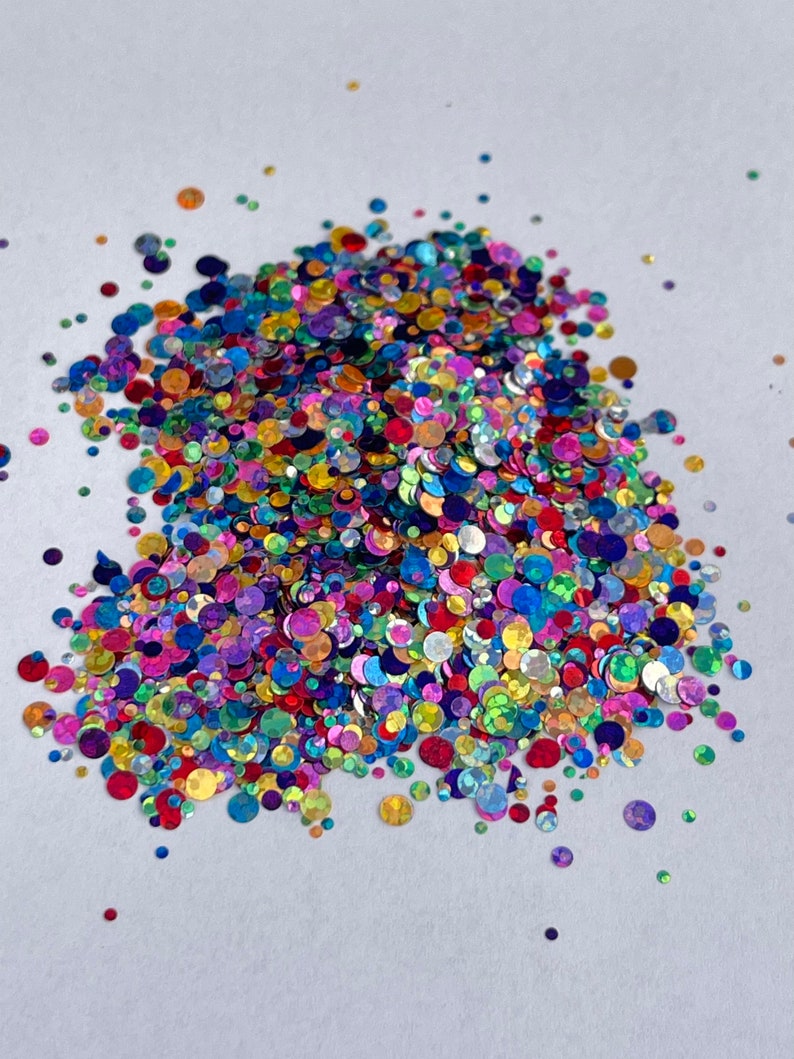 May include: A pile of colorful, iridescent glitter in various sizes and shapes. The glitter includes shades of red, blue, green, purple, and gold. The glitter is scattered on a white surface, creating a sparkling effect.