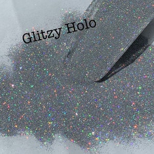 May include: Close-up of a pile of silver glitter with a holographic effect. The glitter is being scooped up by a clear, flat scoop. The text "Glitzy Holo" is printed in black on the white surface.