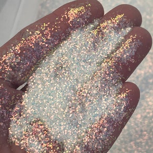 May include: A close-up of iridescent glitter in a hand. The glitter is a mix of white, silver, and gold.