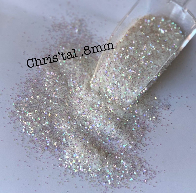 Puede incluir: Primer plano de purpurina iridiscente con una cuchara transparente. La purpurina es una mezcla de blanco y colores brillantes. El texto en la imagen dice "Chris'tal .8mm".