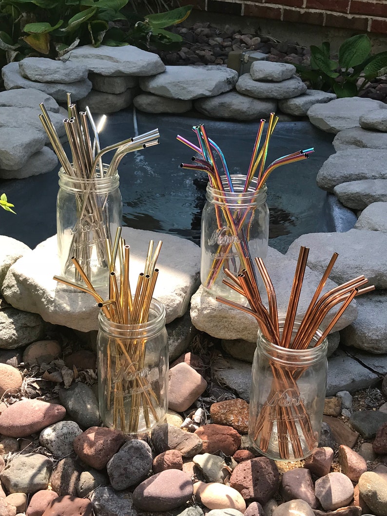 May include: Four glass jars filled with different colored reusable metal drinking straws. The straws are arranged in a circle on a bed of small rocks.