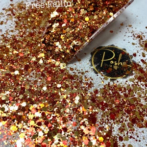 K&ouml;nnte beinhalten: Ein klarer L&ouml;ffel gef&uuml;llt mit Glitzer in Gold-, Rot- und Kupfert&ouml;nen, mit dem Text "Free Fallin'". Der Glitzer ist auf einer wei&szlig;en Oberfl&auml;che verstreut. Ein schwarzes und goldenes Logo ist ebenfalls sichtbar.