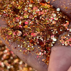 K&ouml;nnte beinhalten: Nahaufnahme einer Hand, die eine Mischung aus Glitzer h&auml;lt. Der Glitzer ist eine Mischung aus Gold-, Bronze- und Rosat&ouml;nen mit verschiedenen Formen und Gr&ouml;&szlig;en. Der Glitzer f&auml;ngt das Licht ein und erzeugt einen funkelnden Effekt.