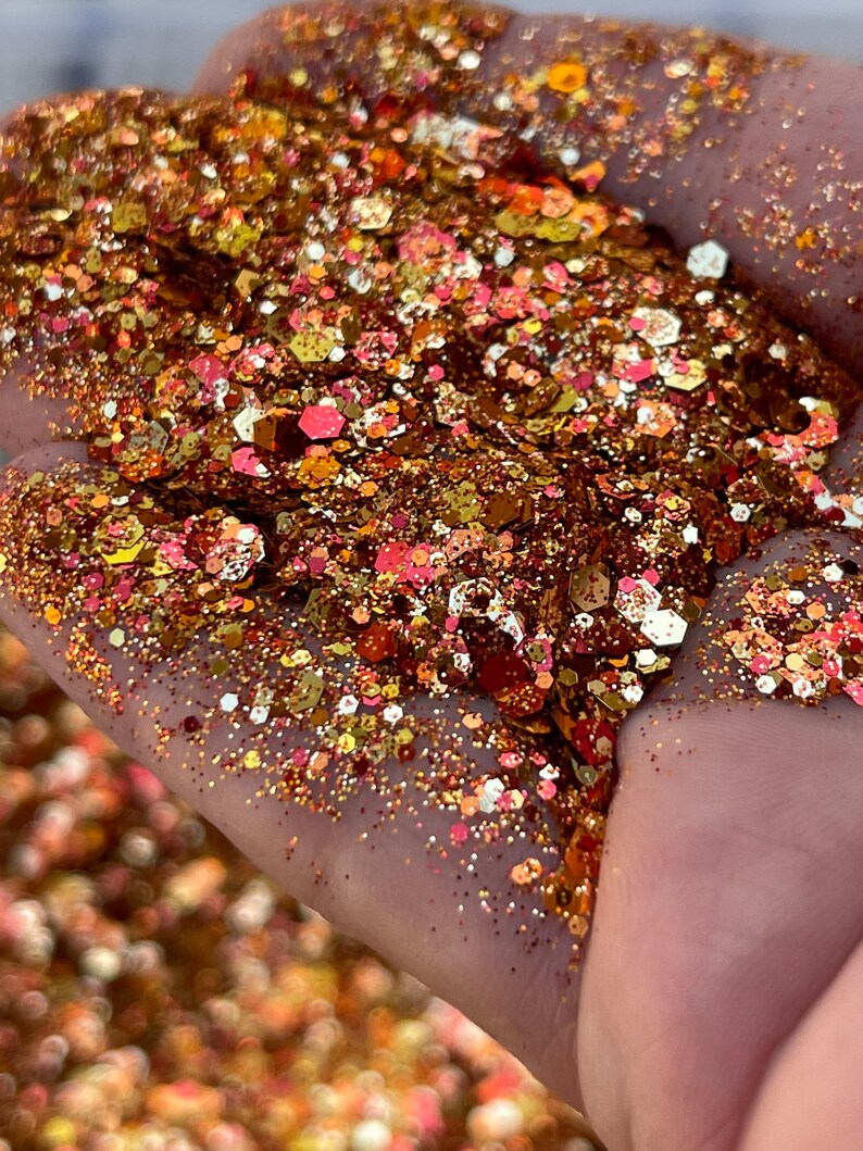 K&ouml;nnte beinhalten: Nahaufnahme einer Hand, die eine Mischung aus Glitzer h&auml;lt. Der Glitzer ist eine Mischung aus Gold-, Bronze- und Rosat&ouml;nen mit verschiedenen Formen und Gr&ouml;&szlig;en. Der Glitzer f&auml;ngt das Licht ein und erzeugt einen funkelnden Effekt.