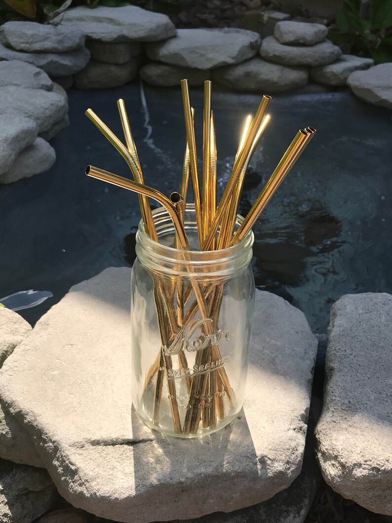 May include: A glass jar filled with a bundle of gold metal drinking straws. The jar is sitting on a gray rock.