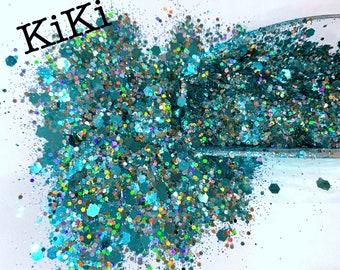 May include: Teal, iridescent glitter with a variety of sizes and shapes. The glitter is scattered on a white surface and the word "Kiki" is written in black at the top of the image.