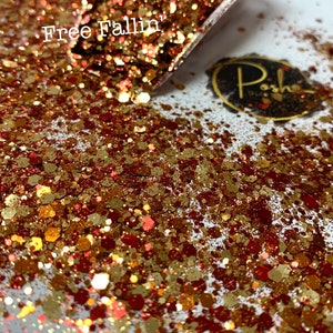 K&ouml;nnte beinhalten: Nahaufnahme eines Glitzermixes in Gold-, Kupfer- und Rott&ouml;nen. Der Glitzer ist auf einer wei&szlig;en Oberfl&auml;che verstreut, wobei sich etwas davon in einer transparenten Schaufel befindet. Der Text "Free Fallin" ist sichtbar. Ein kleines, ovales Logo mit goldenem Rand ist ebenfalls vorhanden.