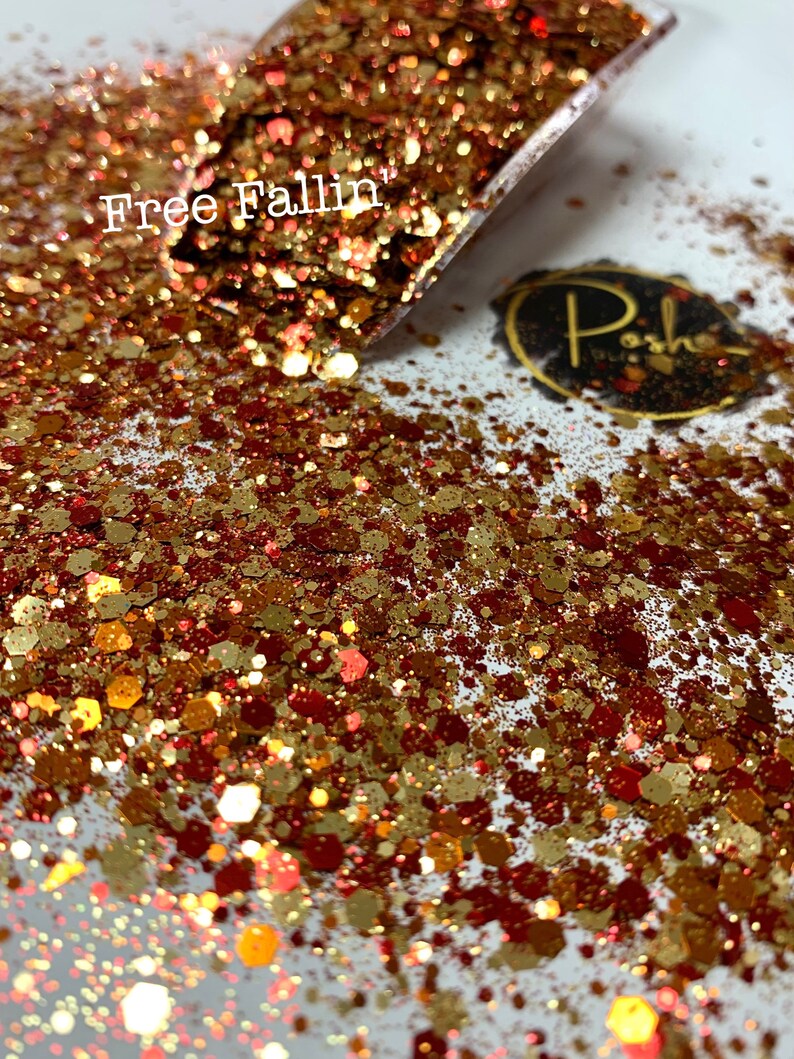 K&ouml;nnte beinhalten: Nahaufnahme eines Glitzermixes in Gold-, Kupfer- und Rott&ouml;nen. Der Glitzer ist auf einer wei&szlig;en Oberfl&auml;che verstreut, wobei sich etwas davon in einer transparenten Schaufel befindet. Der Text "Free Fallin" ist sichtbar. Ein kleines, ovales Logo mit goldenem Rand ist ebenfalls vorhanden.