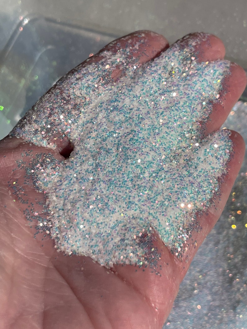 May include: A close-up of iridescent white glitter with a subtle blue shimmer. The glitter is spread out on a hand, showcasing its texture and sparkle.