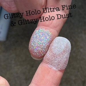 May include: Close-up of two fingers dusted with glitter. One finger displays Glitzy Holo Ultra Fine glitter, and the other shows Glitzy Holo Dust. The glitter reflects multiple colors, creating a shimmering effect. Text in the image reads "Glitzy Holo Ultra Fine & Glitzy Holo Dust."
