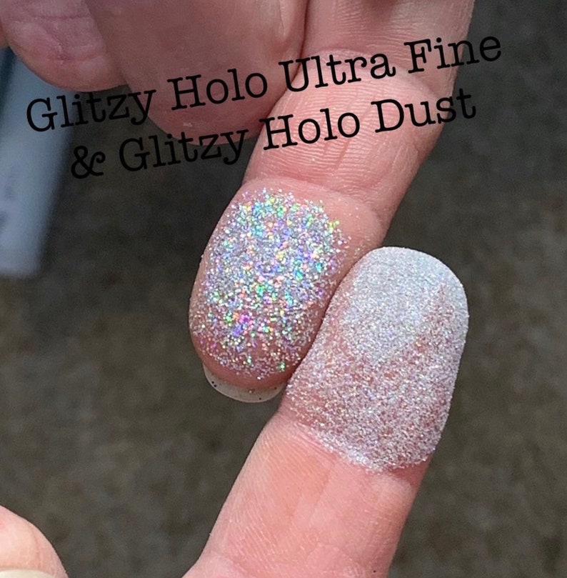 May include: Close-up of two fingers dusted with glitter. One finger displays Glitzy Holo Ultra Fine glitter, and the other shows Glitzy Holo Dust. The glitter reflects multiple colors, creating a shimmering effect. Text in the image reads "Glitzy Holo Ultra Fine & Glitzy Holo Dust."