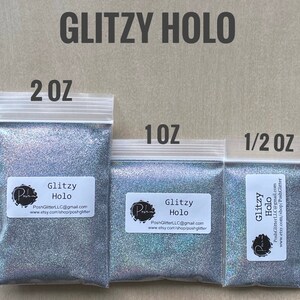 May include: Three clear plastic bags filled with silver glitter. The bags are labeled with the text "GLITZY HOLO" and sizes 2 OZ, 1 OZ, and 1/2 OZ. Each bag has a white label with the brand name and contact information.