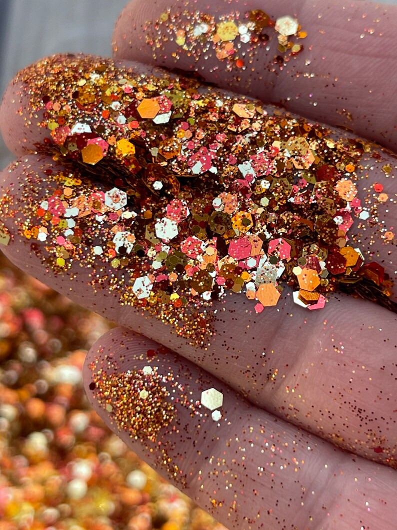K&ouml;nnte beinhalten: Nahaufnahme einer Handvoll Glitzer in Gold-, Kupfer- und Rosat&ouml;nen. Die Glitzerpartikel sind eine Mischung aus kleinen Punkten und gr&ouml;&szlig;eren sechseckigen Formen, die einen funkelnden Effekt erzeugen. Ideal f&uuml;r Bastelarbeiten und Dekorationen.