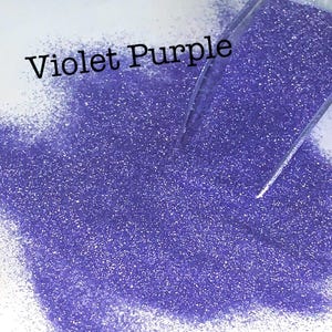 May include: A pile of violet purple glitter is shown, with a clear scoop partially visible on the right side. The words "Violet Purple" are written in black, slightly angled across the top of the glitter.