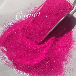 May include: Bright pink glitter spilling from a clear scoop onto a white surface. The word "Cosmo" is written in cursive across the scoop. The glitter is fine and sparkly, creating a vibrant and eye-catching display.