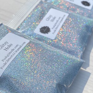 May include: Close-up of three clear plastic bags filled with silver glitter. Each bag has a white label with the text "Glitzy Holo" and a logo. The glitter sparkles with iridescent colors.