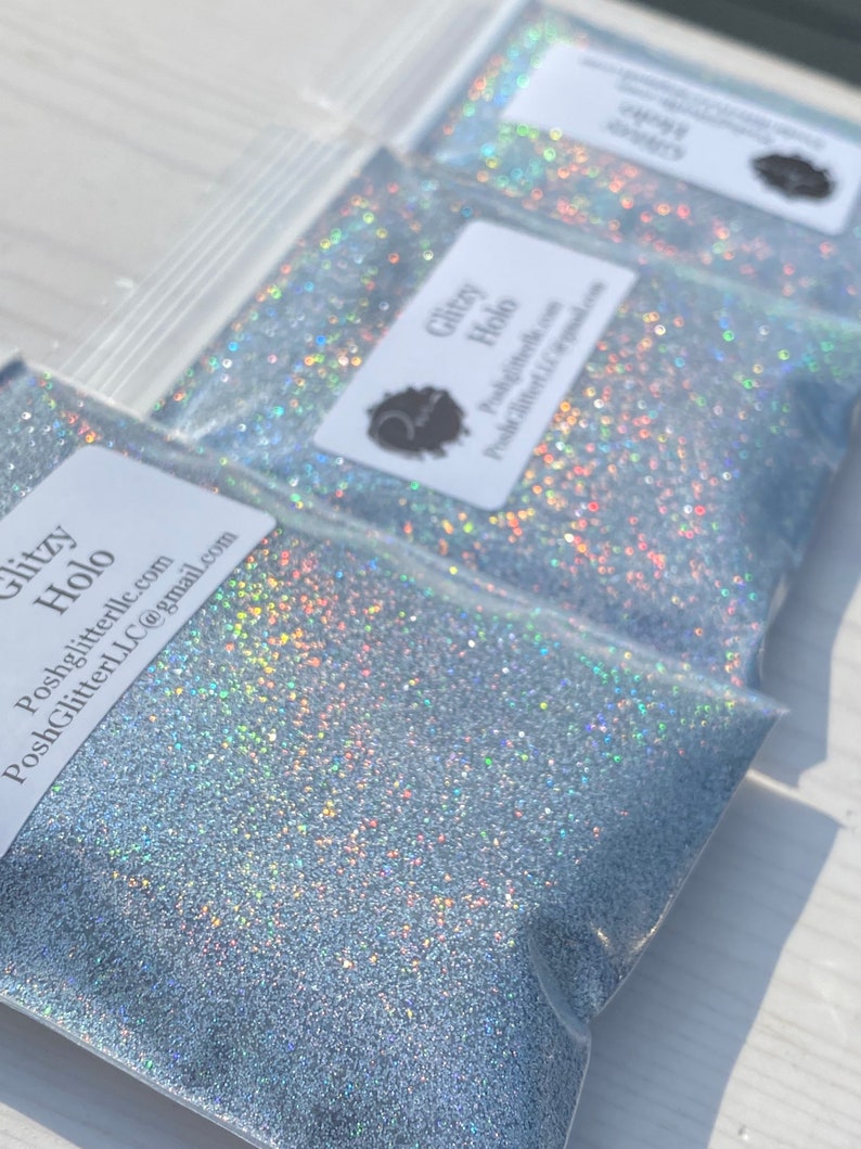 May include: Close-up of three clear plastic bags filled with silver glitter. Each bag has a white label with the text "Glitzy Holo" and a logo. The glitter sparkles with iridescent colors.