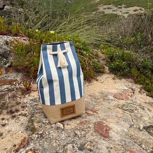 Puede incluir: Una bolsa de lona de rayas azules y blancas con una etiqueta de cuero marrón que dice "Travel Lisboa". La bolsa tiene un asa de cuerda blanca con un borla.