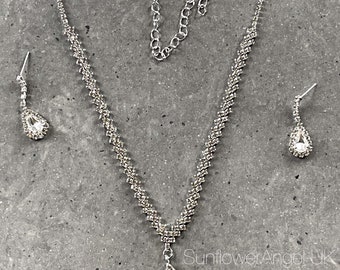 May include: A silver necklace with a teardrop-shaped pendant and matching earrings. The necklace and earrings are adorned with clear rhinestones.