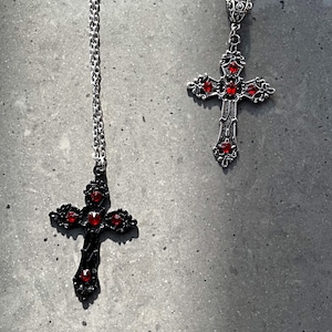 Puede incluir: Dos cadenas de plata con colgantes de cruz negros y plateados. Cada cruz tiene acentos de piedras preciosas rojas. Las cruces son ornamentadas y detalladas. Ash and Co Jewellery está impreso debajo de las cruces.