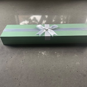 May include: A light green gift box with a white satin ribbon and bow. The box is closed and has a smooth surface.