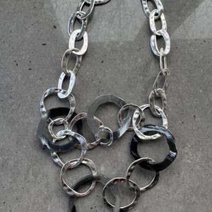 May include: Silver-toned statement necklace featuring a mix of large, textured, and oval-shaped links. The necklace has a cluster of overlapping rings in the center, with some rings having a black finish. The chain is connected with a lobster clasp.