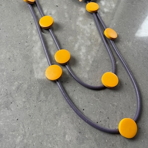 May include: A multi-strand necklace with a purple cord and yellow circular beads. The necklace has a large purple circle at the top.