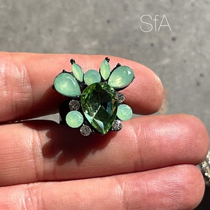 Peut inclure: Une bague noire avec une grande pierre verte en forme de poire entourée de petites pierres vertes et de strass transparents.