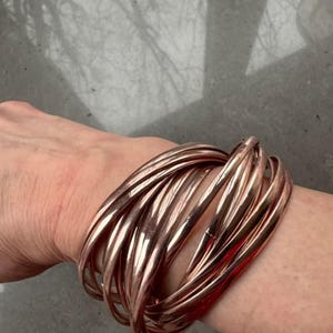 Könnte beinhalten: Ein Set aus roségoldenen Armreifen. Die Armreifen bestehen aus dünnem, abgerundetem Metall und sind an einem Handgelenk gestapelt. Die Armreifen haben eine glänzende, reflektierende Oberfläche. Der Gesamtstil ist modern und minimalistisch.