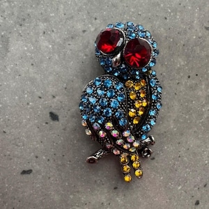 Puede incluir: Broche de búho enjoyado con base de metal plateado. El búho presenta grandes ojos de gemas rojas, plumas de cristal azul y amarillo, y detalles iridiscentes. Un accesorio decorativo para ropa o bolsos.