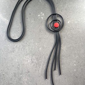 Könnte beinhalten: Eine schwarze Lederkette mit einem runden Anhänger, der einen roten Kreis in der Mitte trägt.