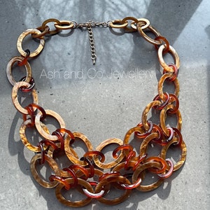 May include: A statement necklace with large, oval links in shades of brown and amber. The necklace has a gold-toned chain and clasp. The text "Ash and Co Jewellery" is visible on the necklace.