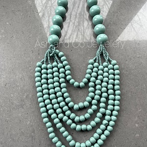 Könnte beinhalten: Eine mehrreihige Halskette aus hellblauen Holzperlen. Die Halskette hat einen Verschluss aus silberner Kette und ist in einem kaskadierenden Stil geschichtet.