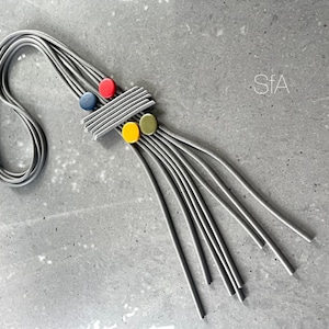 Könnte beinhalten: Eine graue mehrsträngige Halskette mit einem geometrischen Anhänger mit roten, blauen, gelben und grünen Kreisen. Der Anhänger besteht aus grauen und weißen Streifen.
