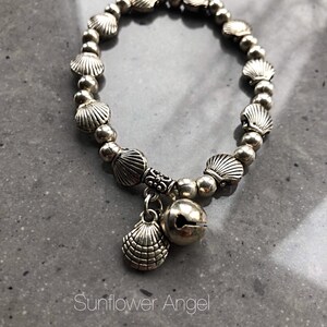 May include: Silver bracelet with a seashell charm and a bell charm. The bracelet is made of small silver beads and seashells.