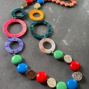 Puede incluir: Un collar llamativo y colorido con cuentas grandes, redondas y ovaladas en varios colores, incluyendo rojo, azul, verde y rosa. El collar también incluye detalles de disco texturizados en tono dorado y una hebra de cuentas color melocotón.