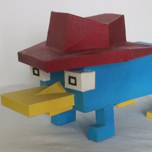 Puede incluir: Una figura de ornitorrinco azul y amarillo con un sombrero rojo. La figura está hecha de cartón y tiene un diseño simple y de bloques.